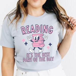 May include: A light blue t-shirt with a pink cartoon stack of books with a smiling face and the text "READING IT'S THE BEST PART OF THE DAY".