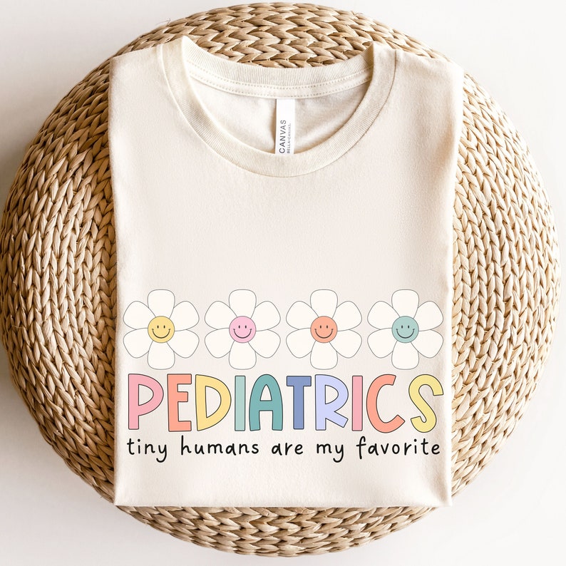 Pu&ograve; includere: Una maglietta color crema con il testo "PEDIATRICS" in colori arcobaleno e la frase "tiny humans are my favorite" sotto. Il testo &egrave; circondato da quattro margherite bianche con centri gialli e facce sorridenti.