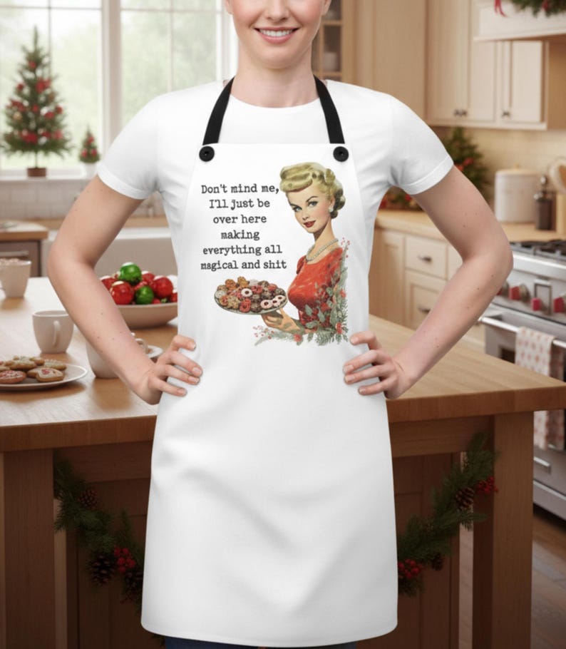 Puede incluir: Delantal blanco con correas negras. Presenta una ilustraci&oacute;n vintage de una mujer sosteniendo un plato de galletas y el texto "Don't mind me, I'll just be over here making everything all magical and shit."