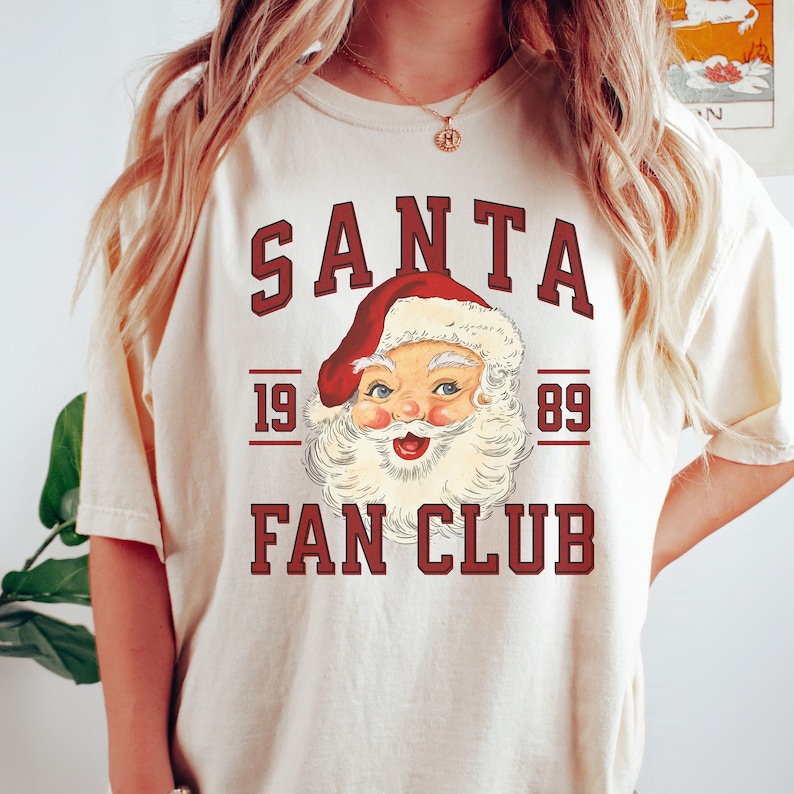 May include: A white t-shirt with a vintage-style graphic of Santa Claus. The graphic says "SANTA FAN CLUB" with the years "19 89" on either side of the Santa image.