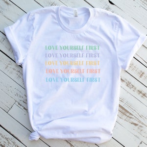 K&ouml;nnte beinhalten: Wei&szlig;es T-Shirt mit dem Spruch "LOVE YOURSELF FIRST", der f&uuml;nfmal in verschiedenen Pastellfarben wiederholt wird.