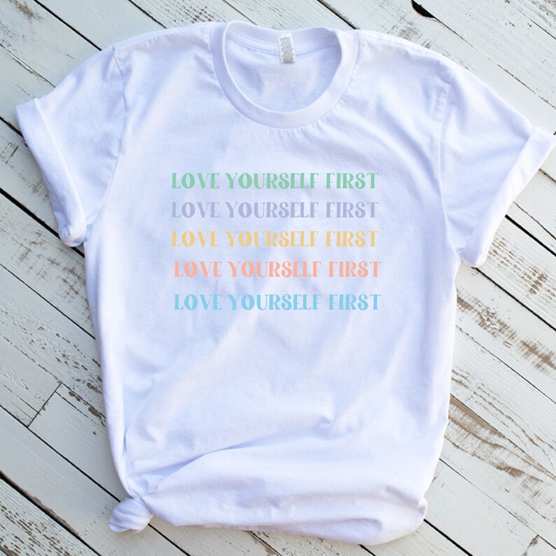 K&ouml;nnte beinhalten: Wei&szlig;es T-Shirt mit dem Spruch "LOVE YOURSELF FIRST", der f&uuml;nfmal in verschiedenen Pastellfarben wiederholt wird.