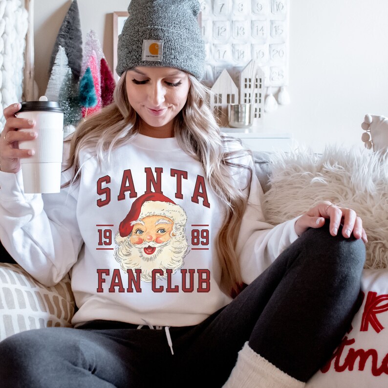May include: White sweatshirt with a red and white Santa Claus graphic and the text "SANTA 19 89 FAN CLUB".