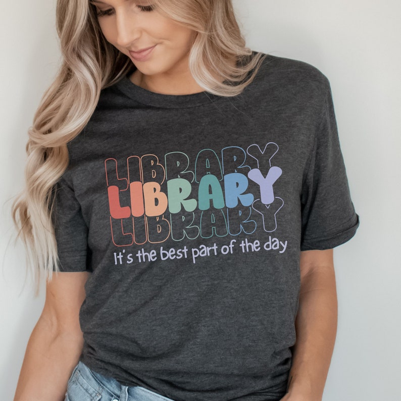 May include: A dark heather grey t-shirt with a rainbow graphic of the word "LIBRARY" repeated three times. The text "It's the best part of the day" is printed below the graphic.
