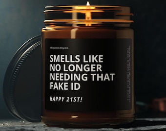 May include: A brown glass candle jar with a black label that reads "SMELLS LIKE NO LONGER NEEDING THAT FAKE ID HAPPY 21ST!". The lid is off to the side. The candle is lit.