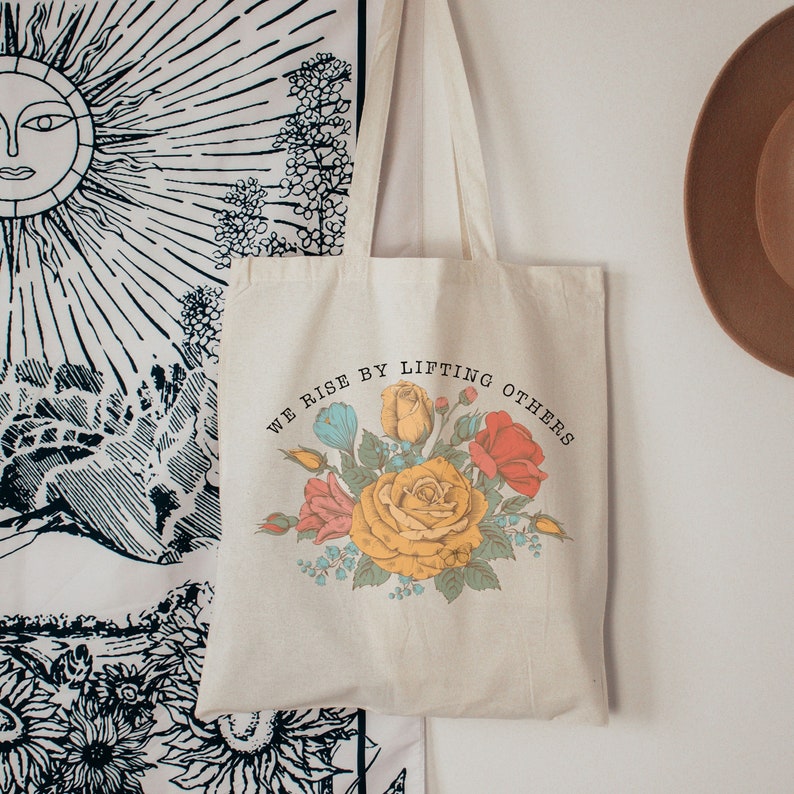 May include: A white canvas tote bag with a floral design and the text "We rise by lifting others".