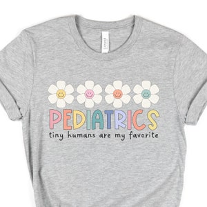 Pu&ograve; includere: Maglietta grigia con una fila di cinque margherite colorate con facce sorridenti e il testo "PEDIATRICS tiny humans are my favorite".