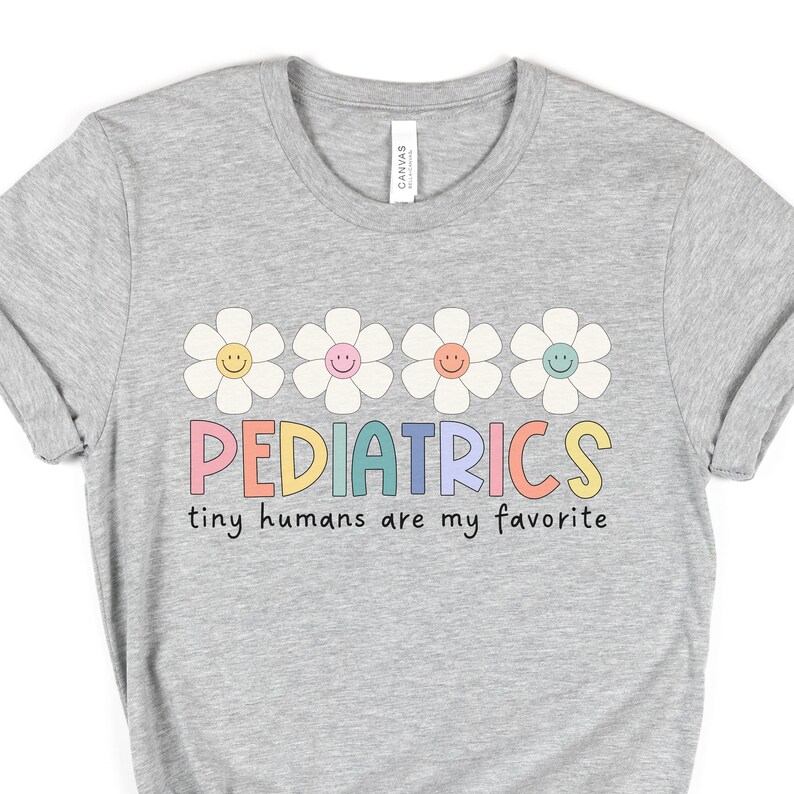 Pu&ograve; includere: Maglietta grigia con una fila di cinque margherite colorate con facce sorridenti e il testo "PEDIATRICS tiny humans are my favorite".