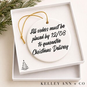 Peut inclure: Une d&eacute;coration ronde en c&eacute;ramique blanche avec un cordon dor&eacute;, portant le texte noir en cursive : All orders must be placed by 12/08 to guarantee Christmas Delivery. Un petit dessin au trait noir d'un sapin de No&euml;l est &eacute;galement visible.