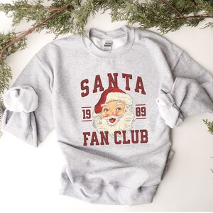 May include: A gray sweatshirt with a red and white Santa Claus graphic and the text "SANTA FAN CLUB 19 89".