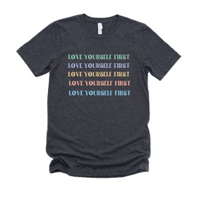 K&ouml;nnte beinhalten: Dunkelgraues T-Shirt mit dem Text "LOVE YOURSELF FIRST", der f&uuml;nfmal in verschiedenen Pastellfarben wiederholt wird.