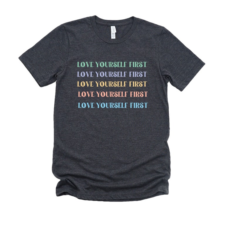 K&ouml;nnte beinhalten: Dunkelgraues T-Shirt mit dem Text "LOVE YOURSELF FIRST", der f&uuml;nfmal in verschiedenen Pastellfarben wiederholt wird.