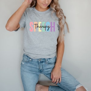 Puede incluir: Una camiseta gris con la palabra "SPEECH" en colores arco&iacute;ris y la palabra "Therapy" escrita en negro debajo.