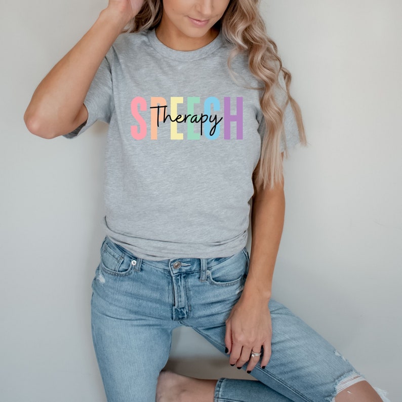Puede incluir: Una camiseta gris con la palabra "SPEECH" en colores arco&iacute;ris y la palabra "Therapy" escrita en negro debajo.