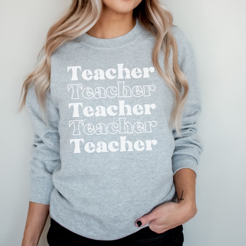 Puede incluir: Una sudadera gris con la palabra "Teacher" repetida cinco veces en una fuente retro blanca.