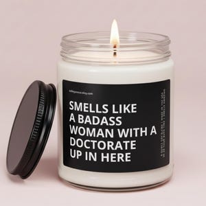 May include: A lit soy wax candle in a clear glass jar with a black lid. The candle has a black label with white text that reads "Smells Like A Badass Woman With A Doctorate Up In Here."