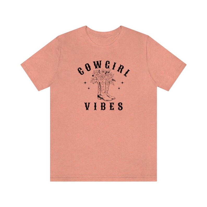 May include: A peach-colored t-shirt with black text that reads "COWGIRL VIBES". A black and white illustration of cowboy boots with flowers in them is above the text.