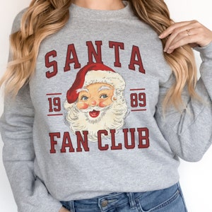 May include: A gray sweatshirt with a vintage-style graphic of Santa Claus. The graphic says "SANTA FAN CLUB" with the years "19 89" on either side of the Santa image.