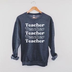 Puede incluir: Una sudadera gris oscuro con la palabra "Teacher" repetida cinco veces en blanco, apilada verticalmente en la parte delantera.