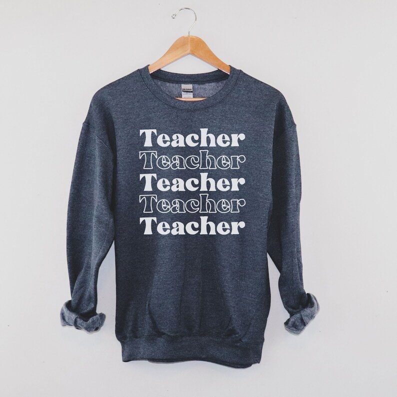 Puede incluir: Una sudadera gris oscuro con la palabra "Teacher" repetida cinco veces en blanco, apilada verticalmente en la parte delantera.