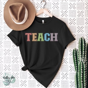 May include: Black t-shirt with the word "TEACH" in rainbow colors. The letters are outlined in white.