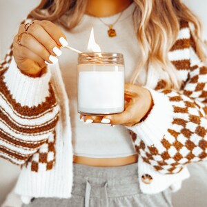K&ouml;nnte beinhalten: Eine brennende Kerze in einem transparenten Glas mit einem wei&szlig;en Etikett. Die Flamme ist hell. Die Person tr&auml;gt eine braun-wei&szlig; karierte Strickjacke und Goldschmuck. Die Kerze befindet sich in einem Glas.
