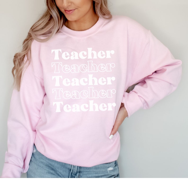 Puede incluir: Una sudadera rosa con la palabra "Teacher" repetida cinco veces en una fuente retro blanca.