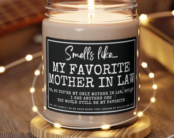 May include: A brown glass candle with a black label that reads "Smells like... My Favorite Mother In Law. Ok, so you're my only mother in law, but if I had another one you would still be my favorite." The candle is sitting on a wooden coaster.