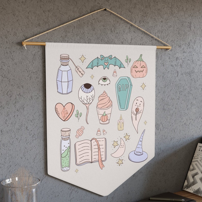 May include: A white canvas banner with a variety of Halloween-themed illustrations, including a bat, a pumpkin, an eye, a heart, a potion bottle, a coffin, a ghost, a book, a witch's hat, and a ghost. The banner has a light brown wooden dowel rod at the top.