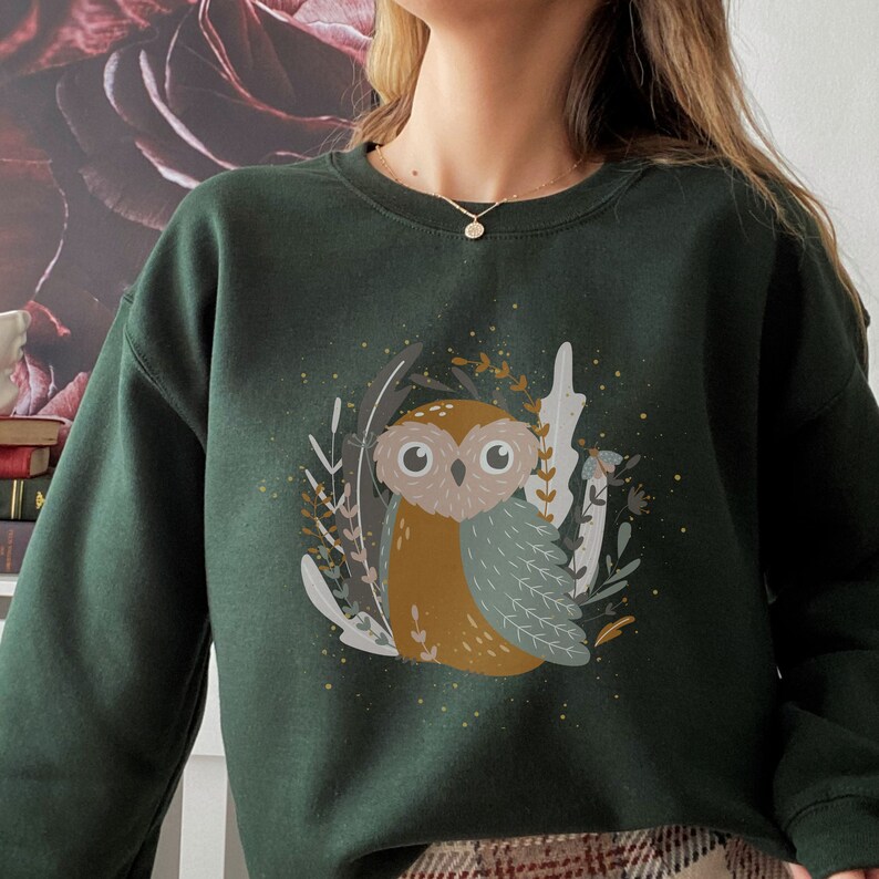 May include: A dark green sweatshirt with a brown and blue owl illustration surrounded by foliage. The owl has large eyes and a whimsical design.