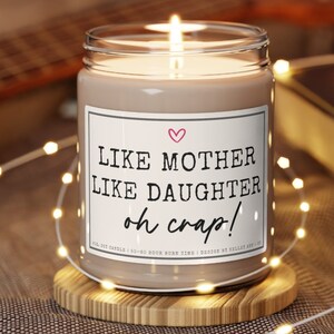 K&ouml;nnte beinhalten: Eine brennende Sojakerze in einem Glas mit einem Etikett mit der Aufschrift "LIKE MOTHER LIKE DAUGHTER oh crap!" mit einem rosa Herz. Die Kerze steht auf einem Holzuntersetzer, umgeben von Lichterketten. Die Kerze hat eine Brenndauer von 50-60 Stunden.
