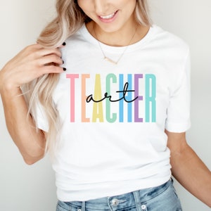 May include: White t-shirt with the words "TEACHER art" in colorful block letters and black cursive. The shirt is a crew neck style and is being worn by a person.