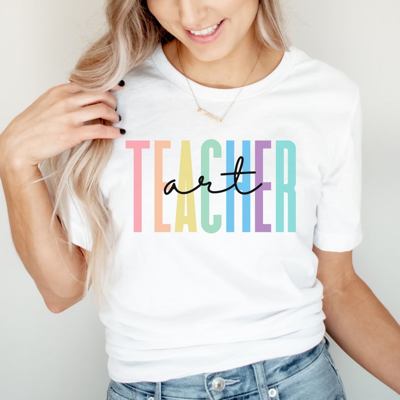 May include: White t-shirt with the words "TEACHER art" in colorful block letters and black cursive. The shirt is a crew neck style and is being worn by a person.