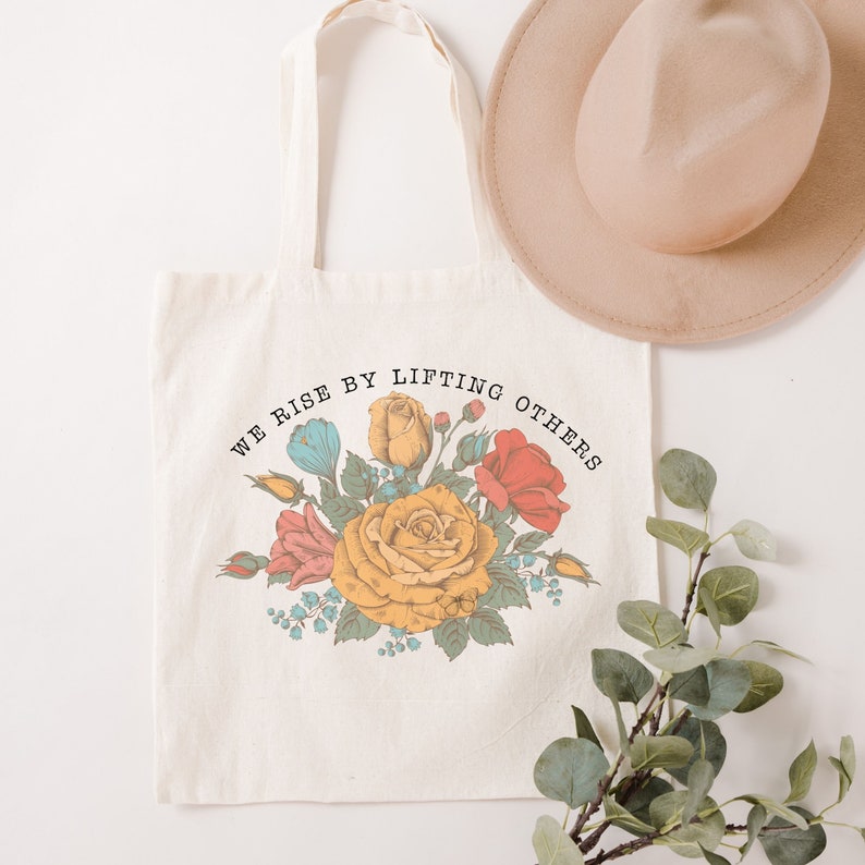 May include: A white canvas tote bag with a floral design and the text "We rise by lifting others".