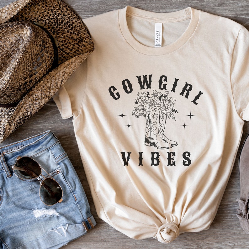 May include: A beige t-shirt with the text "COWGIRL VIBES" in black. The design features a pair of cowboy boots with flowers.