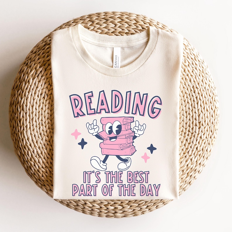 May include: A beige t-shirt featuring a pink and blue graphic of a stack of books with a face and arms. The text "READING IT'S THE BEST PART OF THE DAY" is printed below the graphic.