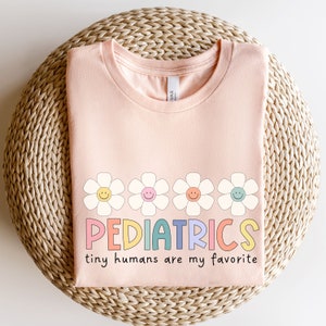 Pu&ograve; includere: Una maglietta rosa con una fila di cinque margherite colorate con facce sorridenti e il testo "PEDIATRICS tiny humans are my favorite".