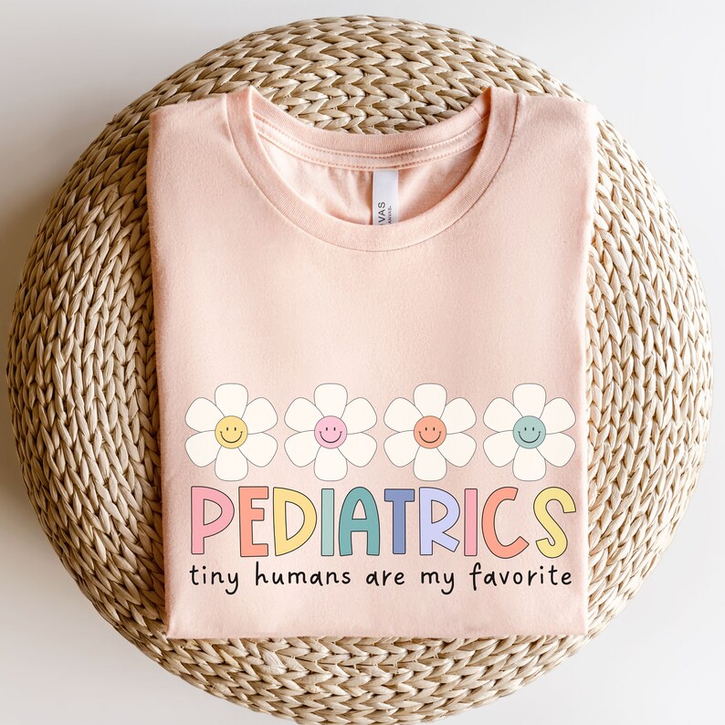 Pu&ograve; includere: Una maglietta rosa con una fila di cinque margherite colorate con facce sorridenti e il testo "PEDIATRICS tiny humans are my favorite".