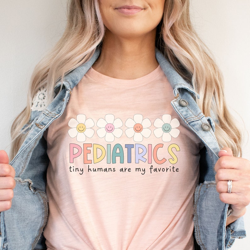 Pu&ograve; includere: Una maglietta rosa con il testo "PEDIATRICS tiny humans are my favorite" in colori arcobaleno. Il testo &egrave; circondato da una fila di cinque margherite bianche con centri gialli e facce sorridenti.