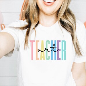 May include: White t-shirt with the word "TEACHER" in pastel colors and the word "art" in a black script font. The shirt is a crew neck style.
