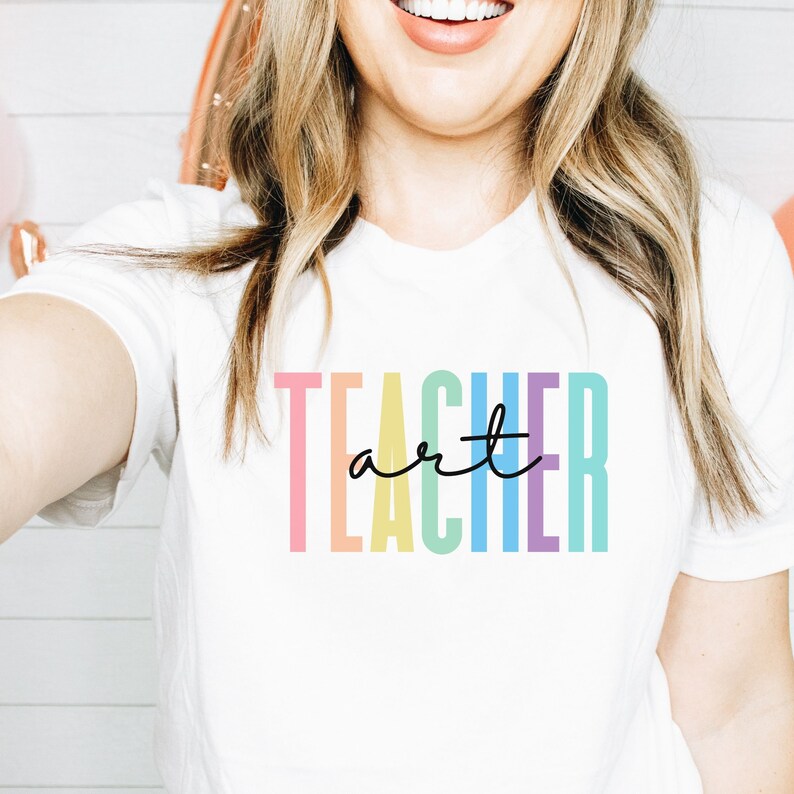 May include: White t-shirt with the word "TEACHER" in pastel colors and the word "art" in a black script font. The shirt is a crew neck style.