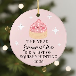 May include: A round, pale pink ceramic ornament featuring a gold hanging string. The center displays a glittery pink dumpling character with a smiling face, surrounded by white stars. Text reads: THE YEAR Samantha DID A LOT OF SQUISHY HUNTING 2026.