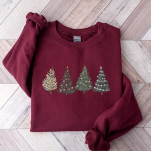 Peut inclure: Sweat-shirt bordeaux avec un motif brod&eacute; de quatre sapins de No&euml;l. Chaque arbre est d&eacute;cor&eacute; de lumi&egrave;res et d'ornements de couleurs diff&eacute;rentes. Les arbres sont dispos&eacute;s en rang&eacute;e sur le devant du sweat-shirt.
