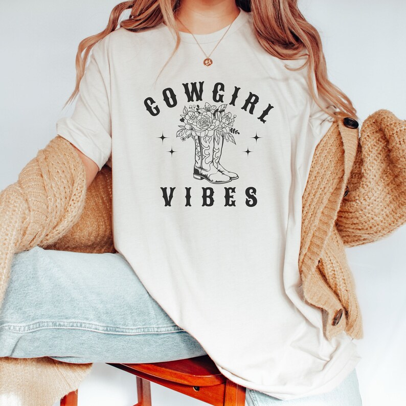 May include: A white t-shirt with a black graphic design of cowboy boots with flowers and the text "COWGIRL VIBES".