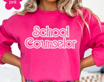 Peut inclure: Un sweat-shirt rose avec le texte "School Counselor" en lettres blanches soulignées.