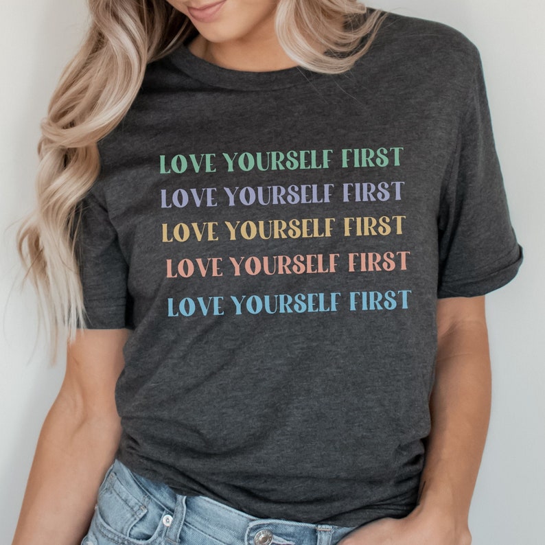 K&ouml;nnte beinhalten: Ein dunkelgraues T-Shirt mit dem Spruch "LOVE YOURSELF FIRST", der f&uuml;nfmal in verschiedenen Pastellfarben wiederholt wird.