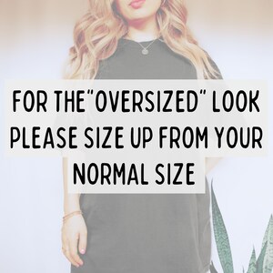 May include: A dark green t-shirt with the text "FOR THE 'OVERSIZED' LOOK PLEASE SIZE UP FROM YOUR NORMAL SIZE" in a white rectangle. The model is wearing a necklace and bracelet.