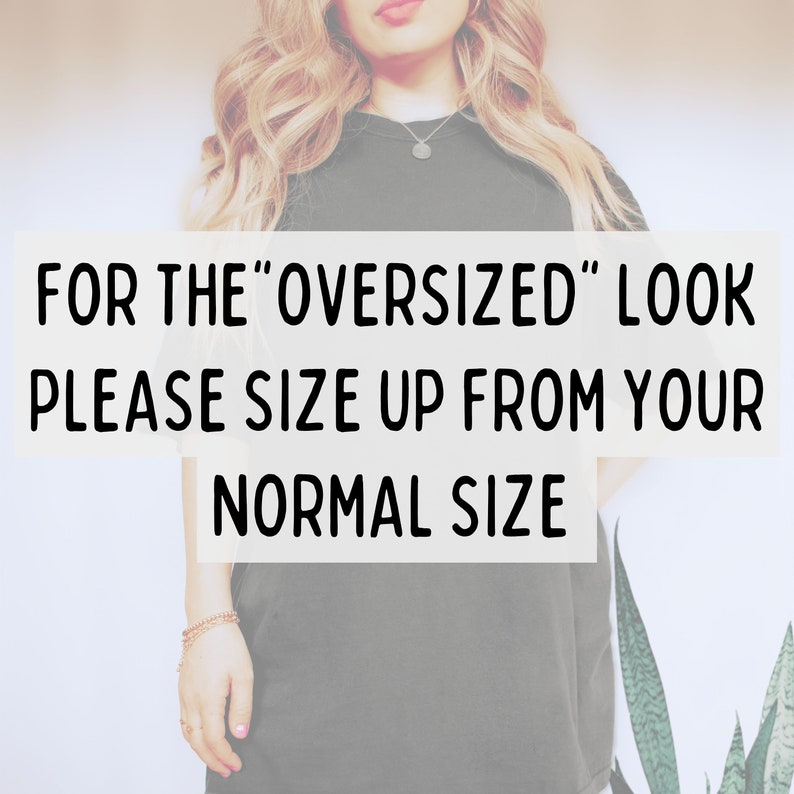 May include: A dark green t-shirt with the text "FOR THE 'OVERSIZED' LOOK PLEASE SIZE UP FROM YOUR NORMAL SIZE" in a white rectangle. The model is wearing a necklace and bracelet.