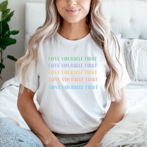 K&ouml;nnte beinhalten: Wei&szlig;es T-Shirt mit dem Spruch "LOVE YOURSELF FIRST", der f&uuml;nfmal in verschiedenen Pastellfarben wiederholt wird.