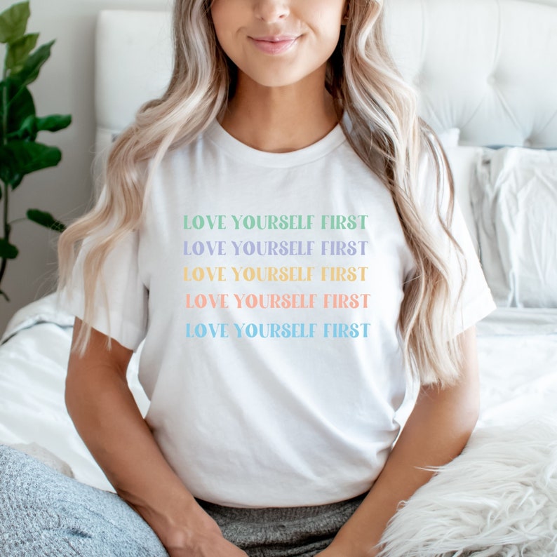 K&ouml;nnte beinhalten: Wei&szlig;es T-Shirt mit dem Spruch "LOVE YOURSELF FIRST", der f&uuml;nfmal in verschiedenen Pastellfarben wiederholt wird.